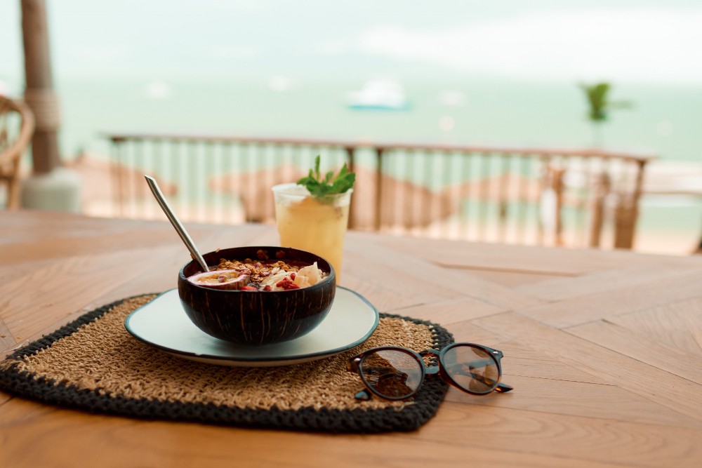 Imagem de uma mesa de restaurante em frente ao mar com prato de comida e óculos de sol.